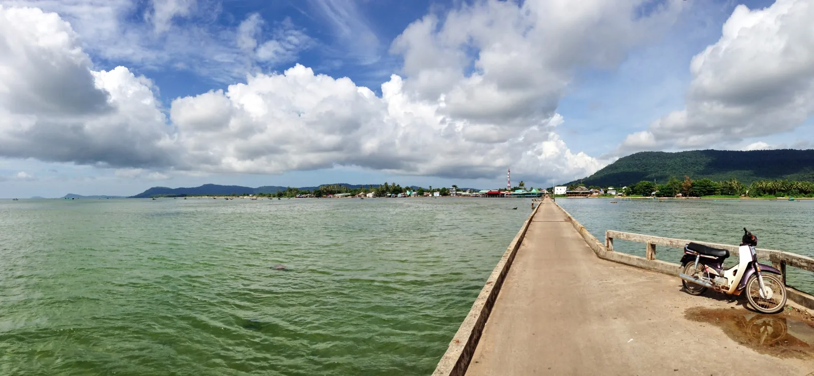 A long concrete pier stretches over green sea water toward a coastal village on Phu Quoc island.