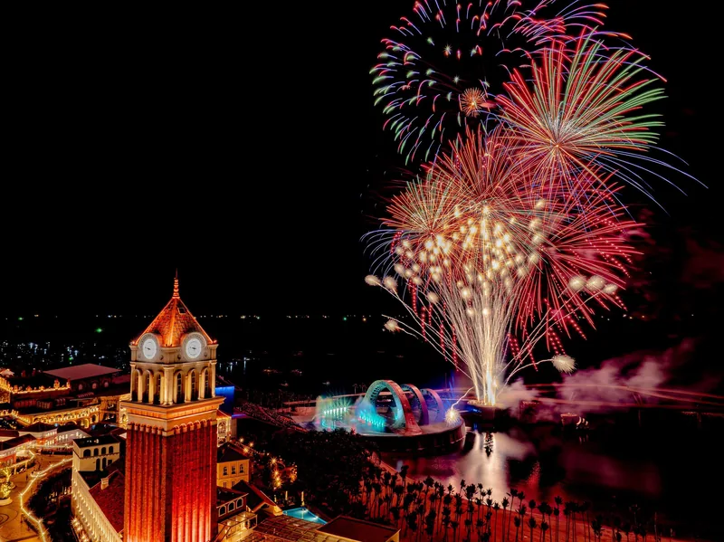 Fireworks burst over a lit clock tower and lagoon at Sunset Town in Phu Quoc at night.
